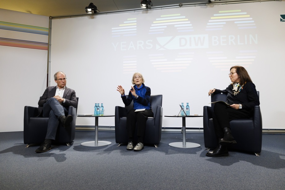Auf dem Podium diskutierten mit Nobelpreisträgerin Claudia Goldin (Mitte): Bernd Fitzenberger, Leiter des Instituts für Arbeitsmarkt- und Berufsforschung (links) und Katharina Wrohlich, Leiterin der Forschungsgruppe Gender Studies am DIW Berlin (rechts); Das Foto zeigt eine Podiumsdiskussion in einem modernen Konferenzraum. Drei Personen sitzen in dunklen Sesseln, die in einem Halbkreis angeordnet sind. Vor jeder Person steht ein kleiner runder Tisch mit mehreren Wasserflaschen darauf. Die mittlere Person gestikuliert mit beiden Händen, während die anderen aufmerksam zuhören. Im Hintergrund ist eine große weiße Leinwand zu sehen, auf der in großen Buchstaben „100 YEARS DIW BERLIN“ steht, wobei die Zahl 100 stilisiert mit farbigen horizontalen Linien dargestellt ist. Die Beleuchtung ist hell und gleichmäßig, und über der Leinwand sind zwei schwarze Scheinwerfer angebracht. Links am Rand des Bildes verläuft ein dekoratives Element mit bunten horizontalen Streifen. Der Boden ist mit einem grauen Teppich bedeckt, und die gesamte Szene wirkt professionell und auf eine formelle Diskussion ausgerichtet.