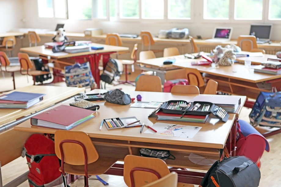 Forschung zu Bildungsungleichheit mithilfe englischer Daten – das hat Bildungsforscher Jascha Dräger für das DIW Berlin gemacht.; Das Foto zeigt ein helles Klassenzimmer mit mehreren Gruppentischen, die jeweils von vier Stühlen umgeben sind. Auf den Tischen liegen verschiedene Schulsachen wie aufgeschlagene Ordner, Mäppchen, Stifte und Papier mit Zeichnungen. Unter den Tischen hängen bunte Schulranzen und Rucksäcke. Im Hintergrund stehen weitere Tische mit Computern und Laptops sowie einige Gegenstände wie Stofftiere und Taschen. Der Raum hat große Fenster, durch die Tageslicht hereinfällt, und der Boden besteht aus hellem Holz. Die Atmosphäre wirkt lebendig und ungeordnet, als wäre der Unterricht gerade unterbrochen oder die Schüler nicht im Raum.