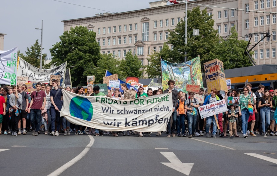 „Wir schwänzen nicht, wir kämpfen“. Die Fridays for Future-Bewegung, hier bei einer Demo am 24. Mai 2019 in Leipzig, habe die gesellschaftliche Stimmung verändert, sagt Karsten Neuhoff.; Das Bild zeigt eine große Gruppe von Menschen, die gemeinsam auf einer Straße vor einem großen, hellen Gebäude stehen und marschieren. Viele von ihnen halten Schilder und Banner hoch, auf denen Forderungen für den Klimaschutz zu lesen sind. Das zentrale Banner vorne ist weiß, zeigt eine Weltkugel und trägt den grünen Schriftzug „FRIDAYS FOR FUTURE. Wir schwänzen nicht, wir kämpfen“. Auch andere Plakate und Transparente sind zu sehen, mit bunten Farben und unterschiedlichen Slogans zum Thema Umweltschutz. Im Hintergrund stehen Bäume und ein gelbes Straßenbahnfahrzeug. Die Stimmung wirkt entschlossen und gemeinschaftlich, und die Menschen setzen sich offenbar für den Schutz des Klimas ein.
