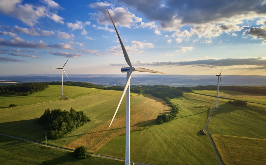 Mit Energie aus Sonne und Wind die Emissionen reduzieren – dafür können Klimaschutzverträge sorgen. Deutschland ist Vorreiter bei diesen Verträgen.; Das Bild zeigt eine weite, leicht hügelige Landschaft mit grünen Feldern und vereinzelten kleinen Wäldern. Im Vordergrund ragt ein großes, weißes Windrad mit drei Flügeln in die Höhe, während im Hintergrund zwei weitere Windräder auf den Feldern verteilt stehen. Am Himmel sind lockere, weiße Wolken zu sehen, durch die Sonnenstrahlen auf die Landschaft fallen. Die Szene wirkt offen, ruhig und vermittelt das Gefühl von Natur und moderner erneuerbarer Energie durch die Windkraftanlagen.