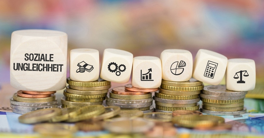 Es gibt einen Zusammenhang zwischen sozialer Ungleichheit und Kriminalität.; Das Foto zeigt mehrere Stapel von Münzen, die in unterschiedlichen Höhen angeordnet sind. Auf den Münzen liegen sechs helle Holzwürfel, von denen jeder ein Symbol trägt. Der erste Würfel trägt die Aufschrift „SOZIALE UNGLEICHHEIT“. Die anderen Würfel zeigen verschiedene Symbole: ein Geldschein mit Münzen, ein Zahnradpaar, ein aufwärts gerichtetes Balkendiagramm, ein Kreisdiagramm, ein Taschenrechner und eine Waage. Die Münzen und Würfel liegen auf einer unscharfen, farbenfrohen Unterlage, die im Hintergrund verschwimmt. Die Szene vermittelt den Eindruck eines visuellen Konzepts zu wirtschaftlichen Themen wie Finanzen, Statistik und sozialer Gerechtigkeit.