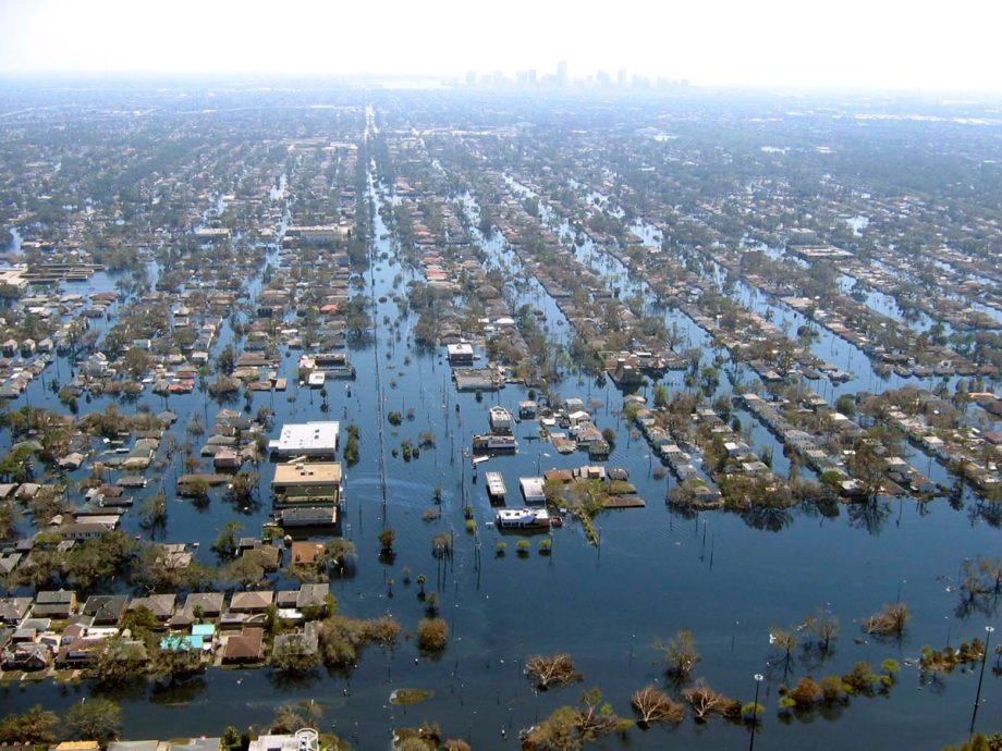 Auch die Wirbelstürmen Katrina und Rita in den USA 2005 hatten Einfluss auf die Rohölpreise.; Das Bild zeigt eine Luftaufnahme einer weitläufigen Stadtlandschaft, die vollständig von Wasser überflutet ist. Die Straßen und Häuser sind deutlich zu erkennen, doch das Wasser steht vielerorts hoch und bedeckt große Bereiche zwischen und um die Gebäude. Die Köpfe von Bäumen und Dächern ragen aus der Flut, während das geordnete Raster der Straßen in der Ausdehnung verschwindet. Im Hintergrund, am Horizont, ist eine entfernte Skyline mit hohen Gebäuden und viel Dunst zu sehen, die die Ausmaße der Überflutung verdeutlicht. Die gesamte Szenerie vermittelt den Eindruck einer verheerenden Flutkatastrophe, bei der das Leben im betroffenen Gebiet nahezu stillzustehen scheint.