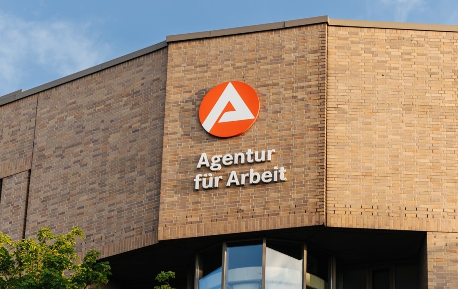 Mit der „Arbeitsmarktdrehscheibe“ will die Arbeitsagentur präventiv arbeiten, erklärt Andrea Nahles beim VfS-Mittagsempfang.; Auf dem Bild ist ein modernes Gebäude aus beigefarbenem Backstein zu sehen. An der Fassade prangt das Logo der „Agentur für Arbeit“, ein roter Kreis mit einem weißen stilisierten A in der Mitte, daneben der Schriftzug „Agentur für Arbeit“ in weißen Buchstaben. Vor dem Gebäude stehen grüne Bäume, und darüber ist ein blauer Himmel mit leichten Wolken zu erkennen.