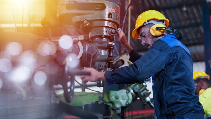 „Ist mein Arbeitsplatz KI-sicher?“. Nahles empfiehlt, dass sich Arbeitnehmer*innen diese Frage stellen. Berufsprofile müssten entsprechend angepasst werden.; Auf dem Bild ist ein Industriearbeiter in einer Werkhalle zu sehen, der an einer großen, schweren Maschine arbeitet. Er trägt einen gelben Schutzhelm, Gehörschutz, Schutzbrille und einen dunkelblauen Arbeitsanzug. Mit beiden Händen bedient er konzentriert die Maschine, während im Hintergrund unscharf weitere technische Geräte und ein zweiter Arbeiter erkennbar sind. Das Licht fällt warm von der Seite ins Bild und erzeugt helle Reflexionen im Vordergrund, die der Szene einen dynamischen und leicht glänzenden Eindruck geben.