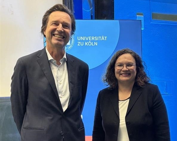 Alexander Kritikos, Vorstandsmitglied im DIW Berlin, moderierte die Veranstaltung mit Andrea Nahles.; Auf dem Bild stehen ein Mann und eine Frau nebeneinander und lächeln in die Kamera. Beide tragen dunkle Blazer über hellen Oberteilen. Im Hintergrund ist ein Schild mit der Aufschrift "Universität zu Köln" zu sehen, das auf einen offiziellen Anlass oder eine Veranstaltung an der Universität hinweist. Die Szenerie wirkt freundlich und professionell, und es handelt sich vermutlich um einen Fototermin bei einer akademischen Veranstaltung.