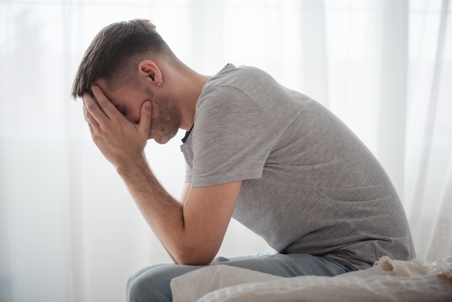 Die psychische Gesundheit junger Menschen hat sich stark verschlechtert.; Das Foto zeigt eine Person, die auf einem Bett oder Sofa sitzt, seitlich zur Kamera gedreht. Die Person trägt ein graues T-Shirt und eine helle Hose. Beide Hände bedecken das Gesicht, die Ellbogen sind auf den Knien abgestützt, der Oberkörper ist leicht nach vorne gebeugt. Der Hintergrund besteht aus hellen, durchscheinenden Vorhängen, die weiches Licht in den Raum lassen.