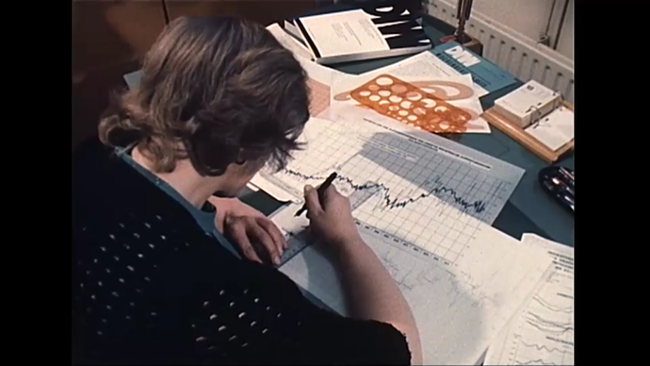 Eine Mitarbeiterin zeichnet eine statistische Abbildung. ; Beschreibung: Das Bild zeigt eine Person, die an einem Schreibtisch arbeitet. Die Person ist konzentriert und scheint Daten oder Diagramme auf einem Blatt Papier zu analysieren.<br />
Visuelle Elemente:<br />
Person: Die Person hat dunkles, lockiges Haar und trägt ein gepunktetes Oberteil.<br />
Sie hält einen Stift in der Hand und arbeitet an einem großen Blatt Papier, das auf dem Schreibtisch liegt.<br />
Schreibtisch: Auf dem Schreibtisch liegen verschiedene Dokumente und Papiere, darunter ein Blatt mit einem Diagramm oder einer Grafik.<br />
Es gibt auch einige Bücher oder Ordner, die auf dem Schreibtisch gestapelt sind.<br />
Ein Lineal und andere Schreibutensilien sind ebenfalls sichtbar.<br />
Hintergrund: Der Hintergrund ist unscharf, was die Aufmerksamkeit auf die Person und ihre Tätigkeit lenkt.<br />
Zusätzliche Informationen: Das Bild vermittelt den Eindruck einer Arbeitsumgebung, möglicherweise in einem Büro oder einer Forschungseinrichtung.<br />
Die Person scheint sich auf eine analytische oder wissenschaftliche Aufgabe zu konzentrieren.<br />
Diese Beschreibung bietet einen Überblick über die Szene einer Person, die an einem Schreibtisch arbeitet und sich auf eine analytische Aufgabe konzentriert.