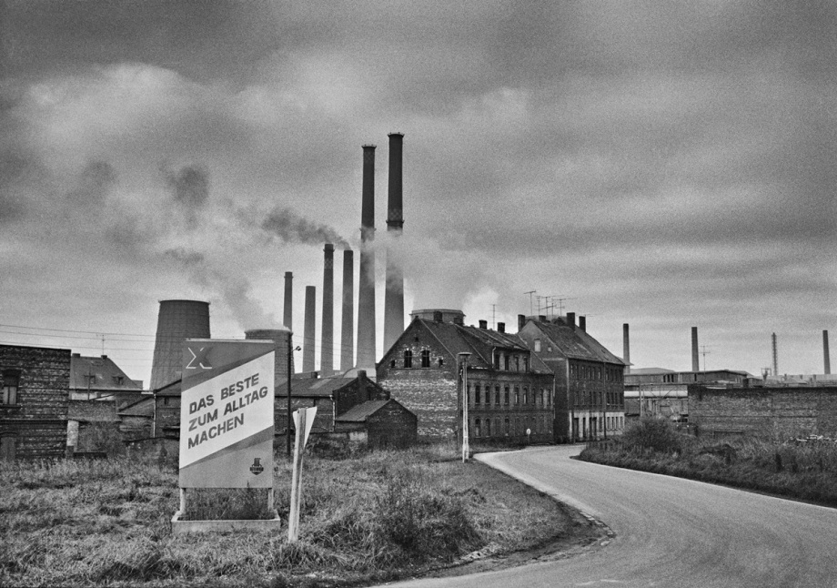 Ein sogenanntes Steinzeugkombinat in Bitterfeld mit rauchenden Schornsteinen im November 1981. ; Bildbeschreibung: Das Schwarzweißfoto zeigt eine Industrielandschaft unter einem stark bewölkten Himmel. Mehrere hohe Schornsteine sind zu sehen, einige stoßen Rauchwolken aus. Im Vordergrund befindet sich ein Schild mit dem Text "DAS BESTE ZUM ALLTAG MACHEN". Das Schild steht in einem überwucherten Grasbereich neben einer Straße, die sich ins Bild hinein windet. Die weiteren Gebäude sehen eher baufällig aus.