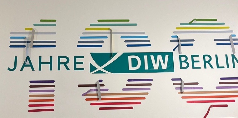 So hatte sich Eva Kretschmer das nicht vorgestellt: Das von ihr gestaltete Logo zu 100 Jahren DIW Berlin löste sich zuerst wieder von der Wand.; Das Foto zeigt eine weiße Wand mit einem grafischen Design. In der Mitte steht in großen grünen Buchstaben „DIW BERLIN“, wobei „DIW“ in einem rechteckigen grünen Feld hervorgehoben ist. Links davon befindet sich das Wort „JAHRE“ in grüner Schrift. Über und unter dieser Schrift sind mehrere horizontale Linien in unterschiedlichen Farben angeordnet, darunter Blau, Grün, Gelb, Lila, Rot und Orange. Die Linien sind in Gruppen angeordnet und wirken wie stilisierte Balkendiagramme oder abstrakte Muster. Einige der sind unterbrochen. Es sieht aus, als hätten sich die Linien des Logos von der Wand gelöst.