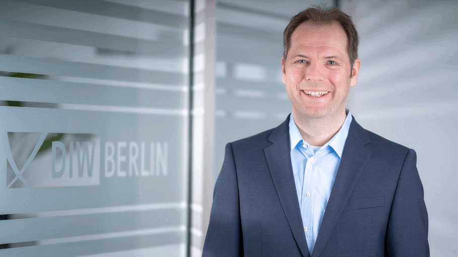 Die Abbildung zeigt DIW-Forscher Hannes Ullrich (Abteilung Unternehmen und Märkte) in einer Innenraumaufnahme. Er steht rechts im Bild und ist vom Oberkörper aufwärts zu sehen. Hannes Ullrich trägt ein dunkelblaues Sakko über einem hellblauen Hemd. Das Gesicht ist zur Kamera ausgerichtet. Im linken Bildbereich befindet sich eine Glaswand mit mehreren horizontalen, mattierten Streifen. Auf der Glaswand ist das Logo „DIW BERLIN“ sichtbar. Der Hintergrund zeigt einen hellen, modernen Büro- oder Institutsflur mit unscharfer Darstellung. Die Beleuchtung ist gleichmäßig und hell. Die Farbgestaltung wird von Grau-, Blau- und Weißtönen geprägt. Weitere Personen oder Gegenstände sind nicht zu sehen. Die Aufnahme vermittelt den Charakter eines professionellen Porträtfotos in einem institutionellen Arbeitsumfeld.