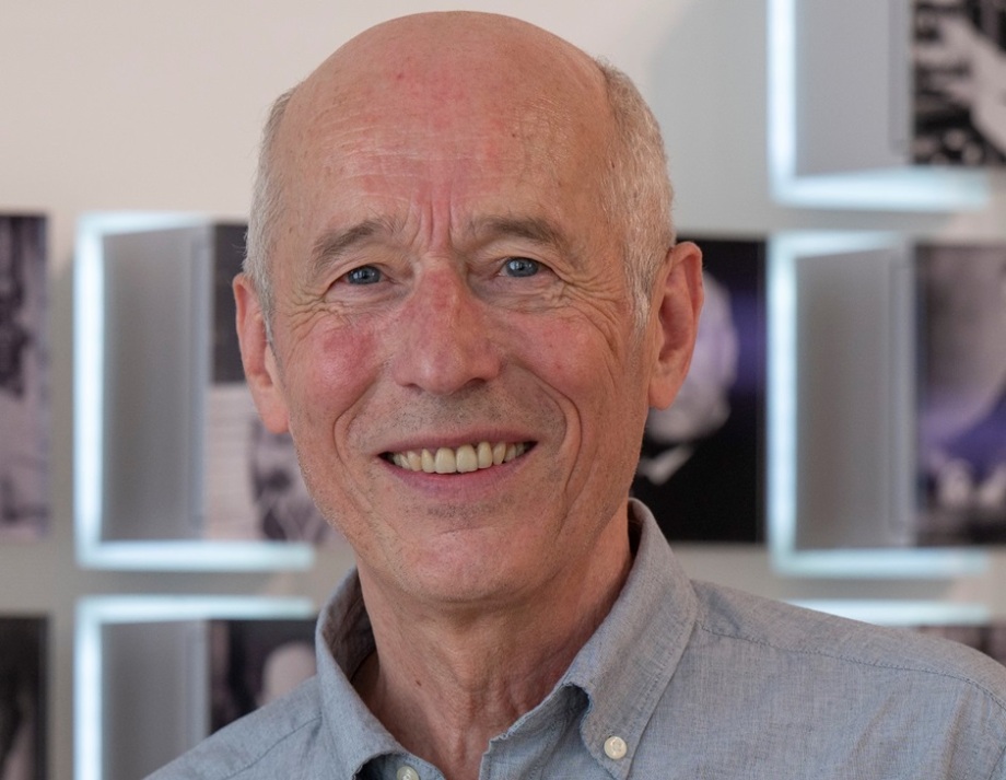 Jürgen Gehards im Juni 2022.; Auf dem Bild ist Jürgen Gerhards zu sehen. Er trägt ein hellblaues Hemd mit Kragen und hat nur wenig graues Haar, besonders am Hinterkopf. Im Hintergrund befinden sich mehrere unscharfe Schwarz-Weiß-Fotografien an einer hellen Wand, die von rechteckigen Leuchtrahmen umgeben sind. Jürgen Gerhards steht im Vordergrund und nimmt den größten Teil des Bildes ein.