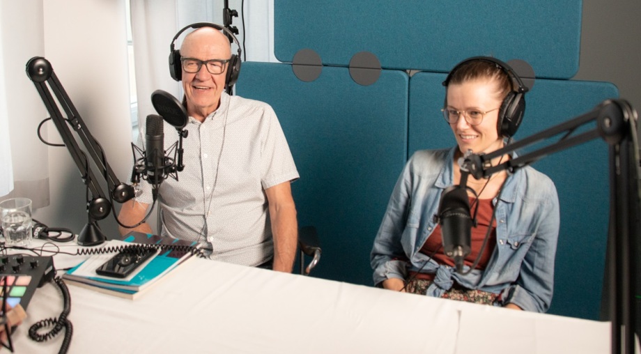 Martin Gornig und Laura Pagenhardt im bei der Podcastaufnahme für „Wirtschaft bewegt – 100 Jahre DIW Berlin“; Zwei Personen, ein Mann mit Glatze und Brille links und eine junge Frau mit Brille rechts, sitzen nebeneinander in einem Podcast-Studio. Sie tragen große Kopfhörer und sprechen jeweils in ein Mikrofon, das auf einem beweglichen Ständer vor ihnen montiert ist. Vor ihnen steht ein Tisch, auf dem mehrere technische Geräte, Bücher, ein Glas Wasser und Kabel liegen. Im Hintergrund ist eine schallgedämmte Wand mit blauen, rechteckigen und runden Elementen zu sehen. Die Atmosphäre wirkt ruhig und konzentriert, typisch für eine Aufnahmeumgebung.