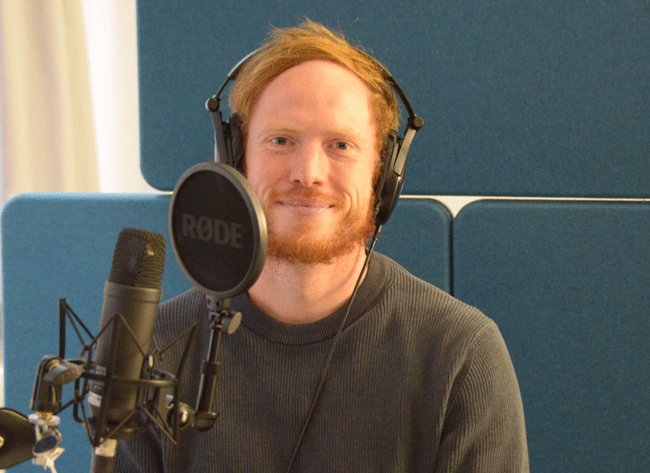 Bildungsforscher Jascha Dräger bei der Aufzeichnung des Podcasts „Wirtschaft bewegt“.; Das Foto zeigt eine Person, die vor einem professionellen Mikrofon sitzt und Kopfhörer trägt. Das Mikrofon ist auf einem Stativ befestigt und hat einen runden Pop-Schutz mit der Aufschrift „RODE“. Die Person trägt einen schlichten, langärmeligen Pullover. Im Hintergrund sind gepolsterte, blau bezogene Schallschutzwände zu sehen, die den Raum akustisch optimieren. Die Beleuchtung ist weich und gleichmäßig, was auf eine Innenaufnahme in einem Studio hinweist. Die Szene vermittelt den Eindruck einer Aufnahme für einen Podcast.