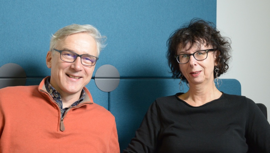 DIW-Finanzexpertin Kristina van Deuverden (rechts) im Gespräch mit Podcasthost Erich Wittenberg.; Auf dem Foto sitzen zwei Menschen nebeneinander vor einer blauen gepolsterten Wand, als wären sie in einer gemütlichen Sitzecke. Links im Bild ist ein Mann mittleren bis höheren Alters mit kurzen grauen Haaren und Brille zu sehen; er trägt einen orangefarbenen Pullover mit Reißverschluss am Kragen, lächelt und wirkt entspannt. Rechts neben ihm sitzt eine Frau ähnlichen Alters mit lockigen dunklen, leicht zerzausten Haaren und Brille; sie trägt ein schlichtes schwarzes Oberteil und auffällige lange Ohrringe und schaut ruhig in die Kamera. Die Szene ist gleichmäßig ausgeleuchtet, der Hintergrund wirkt modern und ruhig, sodass die beiden Personen im Mittelpunkt des Bildes stehen.