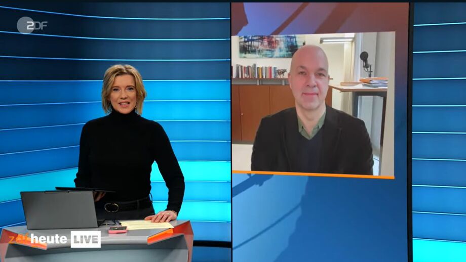 Die Nachrichtensendung ZDF Live wird gezeigt. Links steht die Moderatorin hinter einem Pult in einem modernen Studio mit horizontalen, türkis-blauen Lichtstreifen an der Wand. Auf dem Pult liegen ein aufgeklapptes Laptop, ein Notizblock und einige Stifte. Unten links ist eine Sendungsbauchbinde mit der Aufschrift „ZDF heute LIVE“.<br />
Rechts im Bild befindet sich ein groß eingeblendetes Videofenster. Darin sitzt DIW-Präsident Marcel Fratzscher in einem Büroraum: im Hintergrund sind ein Bücherregal, eine Kommode, ein Tisch mit Papierstapeln sowie ein Mikrofonarm sichtbar. Das Videofenster ist mit einem orangefarbenen Rahmen unten betont.<br />
Komposition & Farben: Linke Bildhälfte: Studio in kühlen Blau- und Türkistönen; schwarze Kleidung der Moderatorin kontrastiert mit der hellen Studiowand.<br />
Rechte Bildhälfte: Videofenster mit warmen Holz- und Beigetönen; orange Akzentlinie am unteren Rand.<br />
Klare Zweiteilung: Studio (links) und Gesprächseinblendung (rechts).<br />
Text/Einblendungen: Logo oben links: „ZDF“.<br />
Bauchbinde unten links: „ZDF heute LIVE“.<br />
Kontext & Funktion: Das Bild zeigt eine Live-Nachrichtensituation mit einer Studiomoderation und einer zugeschalteten Interviewperson. Die Gestaltung unterstützt die Trennung von Moderation (Studio) und Gespräch (Videofenster).<br />
Hinweise zur Zugänglichkeit (gestaltungsbezogen):<br />
Hoher Kontrast zwischen Studiohintergrund und Vordergrundobjekten.<br />
Klare visuelle Hierarchie: Moderatorin links, Interviewfenster rechts, statische Bauchbinde unten links.<br />
Lesbarkeit: kurze, gut platzierte Textelemente ohne Überlagerungen.