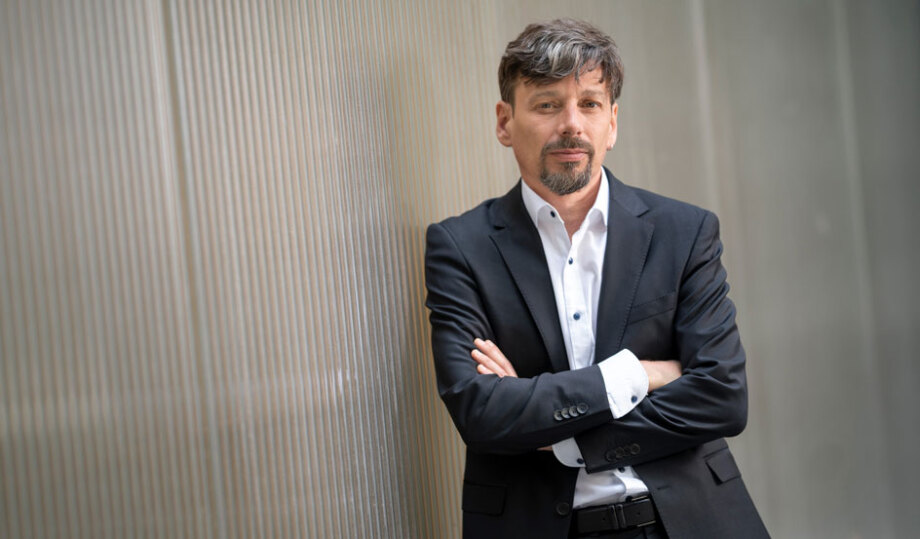 Auf dem Bild ist Tomaso Duso, Leiter der Abteilung Unternehmen und Märkte, zu sehen. Duso steht im Vordergrund vor einer hellen, senkrecht strukturierten Wand. Er trägt ein dunkles Sakko über einem weißen Hemd und hat die Arme vor dem Körper verschränkt. Der Oberkörper ist leicht seitlich ausgerichtet, während das Gesicht zur Kamera zeigt. Die Haare sind kurz und leicht gewellt, das Gesicht ist teilweise von Bart bedeckt. Der Hintergrund besteht aus einer hellen Wand mit feinen vertikalen Linien. Die Beleuchtung ist weich und gleichmäßig, ohne starke Schatten. Es sind keine weiteren Gegenstände oder Texte im Bild sichtbar. Die dunkle Kleidung steht im Kontrast zum hellen Hintergrund.