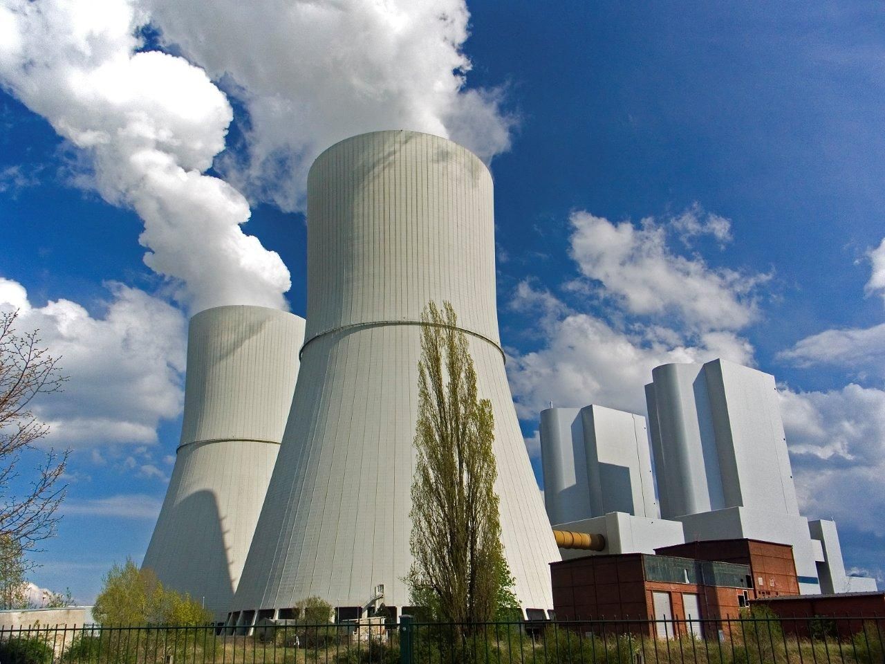 Auf dem Bild ist ein Kraftwerk zu sehen, das durch die Kühltürme starke, weiße Dampfwolken in den blauen Himmel bläst.